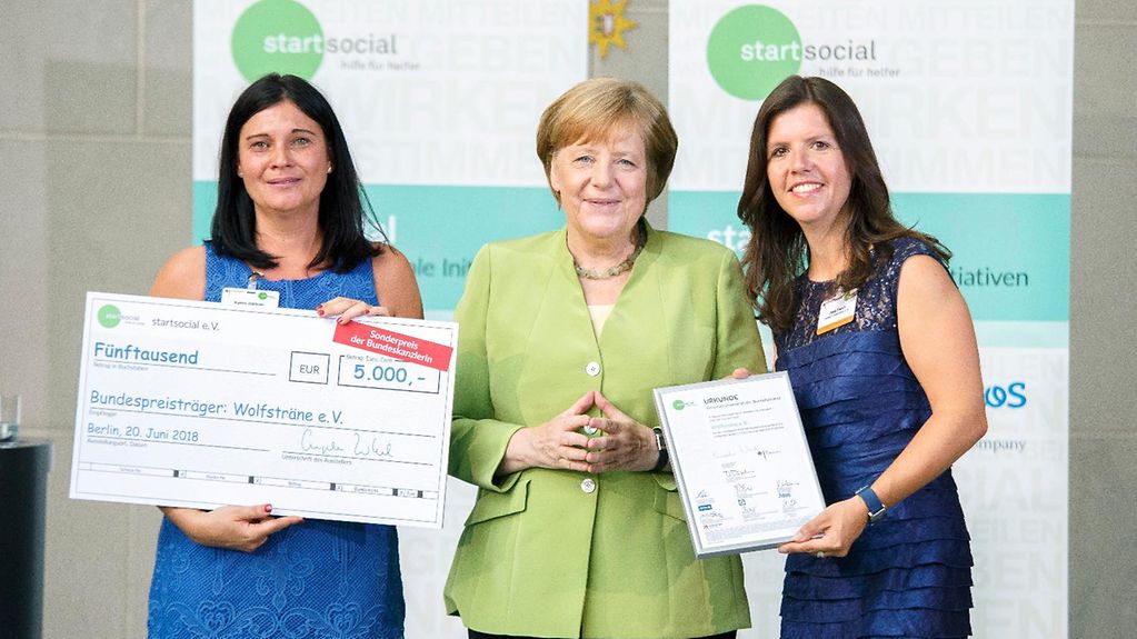 Angela Merkel steh zwischen zwei Frauen in blauen Kleidern. Links hält Karin Gärtner einen Scheck über 5.000 Euro in der Hand. Rechts hält Julia Enoch die Startsocial-Urkunde.
