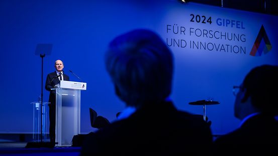 Federal Chancellor Scholz stands on a stage at the 2024 Research Summit where he is giving a speech. The photo is taken from the audience, so the silhouettes of two audience members can be seen.