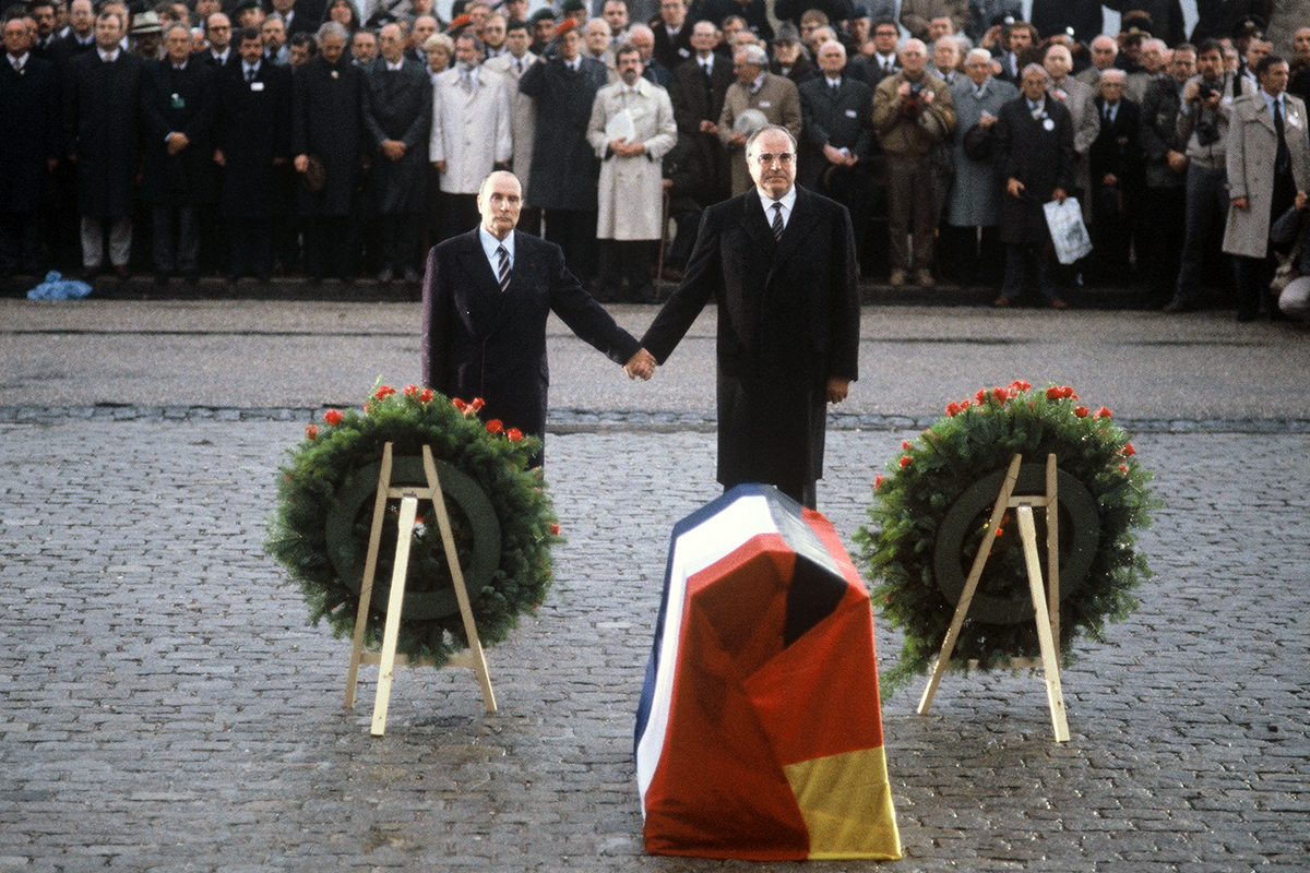 Bundeskanzler Helmut Kohl und der französische Präsident François Mitterrand stehen Hand in Hand vor Trauerkränzen.