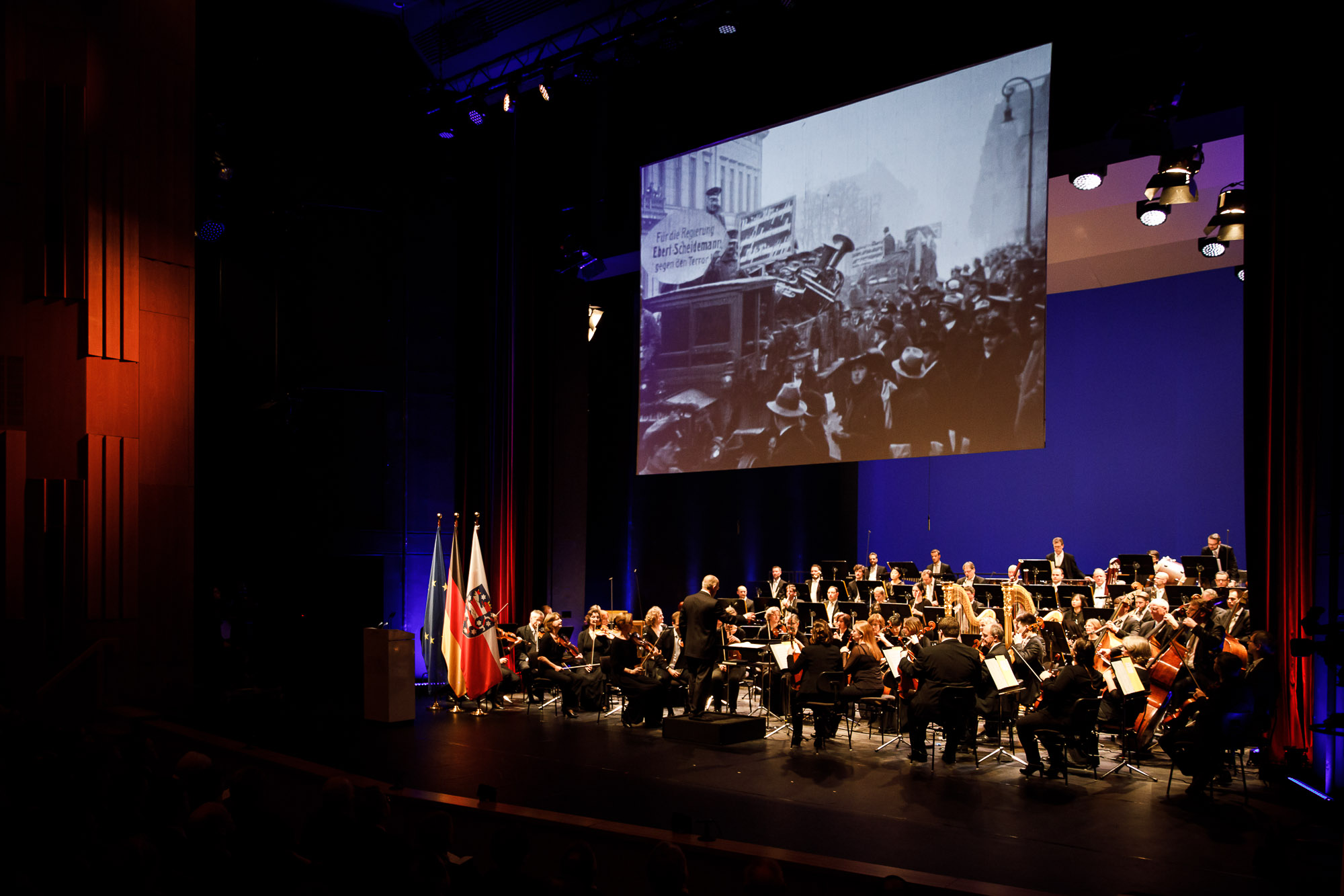 A view of the stage; an orchestra plays while a film from the period is shown on a screen.