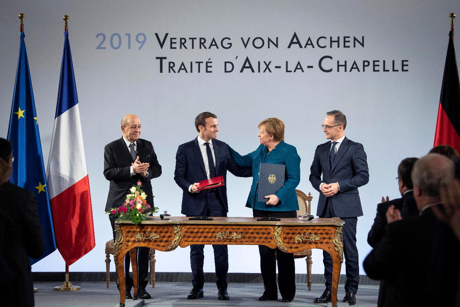 President Emmanuel Macron and Chancellor Angela Merkel at a table, behind them on the wall "2019 Treaty of Aachen". They are flanked by the German and French foreign affairs ministers Heiko Maas and Jean-Yves Le Drian. 