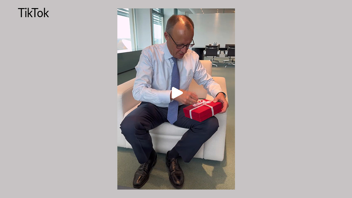 Das Bildschirmfoto zeigt Bundeskanzler Merz mit einem roten Geschenk in der Hand in seinem Büro im Bundeskanzleramt.