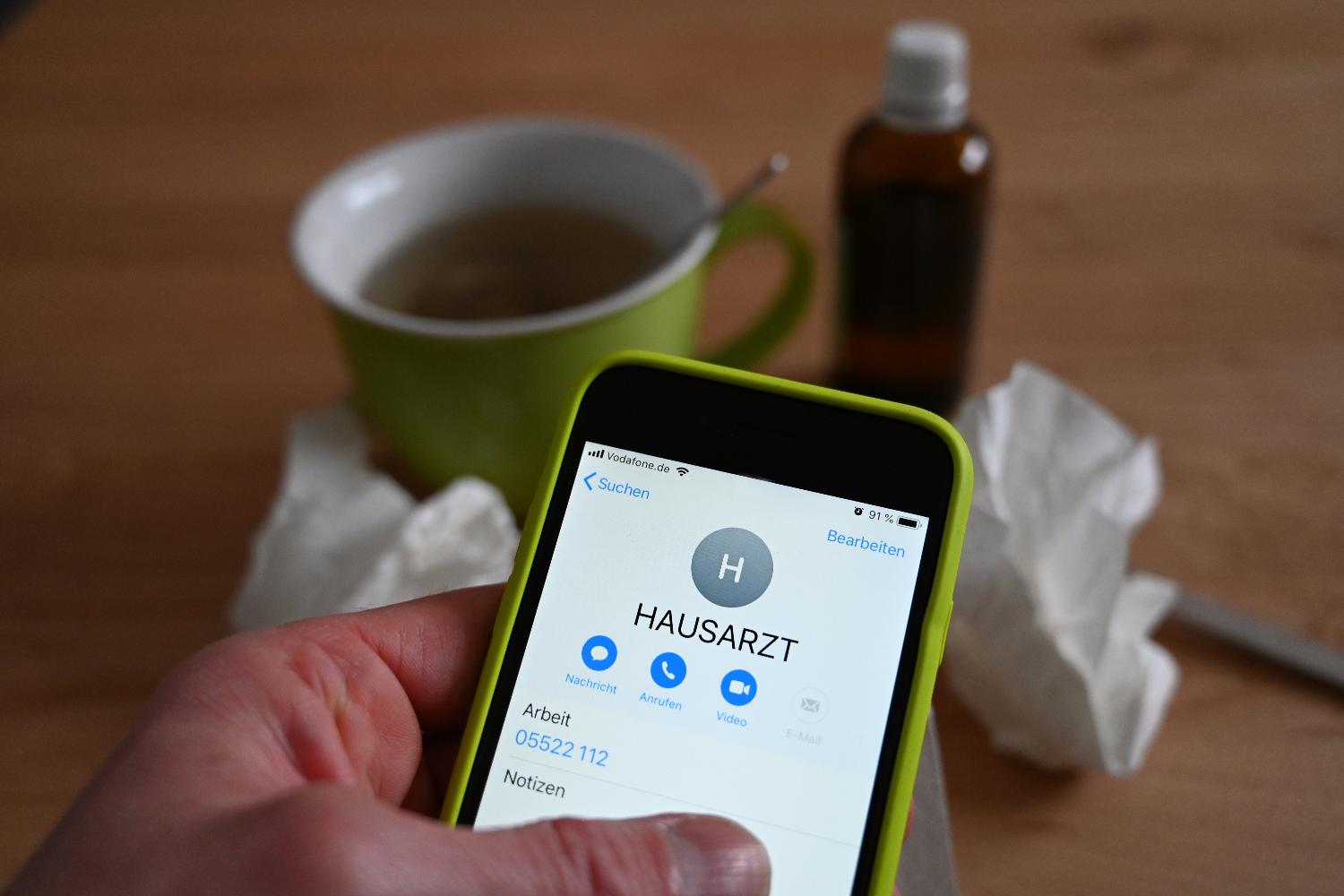 Eine Hand hält ein Handy in der Hand. Auf dem Bildschirm steht „Hausarzt“. Im Hintergrund steht eine grüne Tasse Tee auf einem Tisch. Daneben sind Papiertaschentücher und eine Flasche Medizin. 