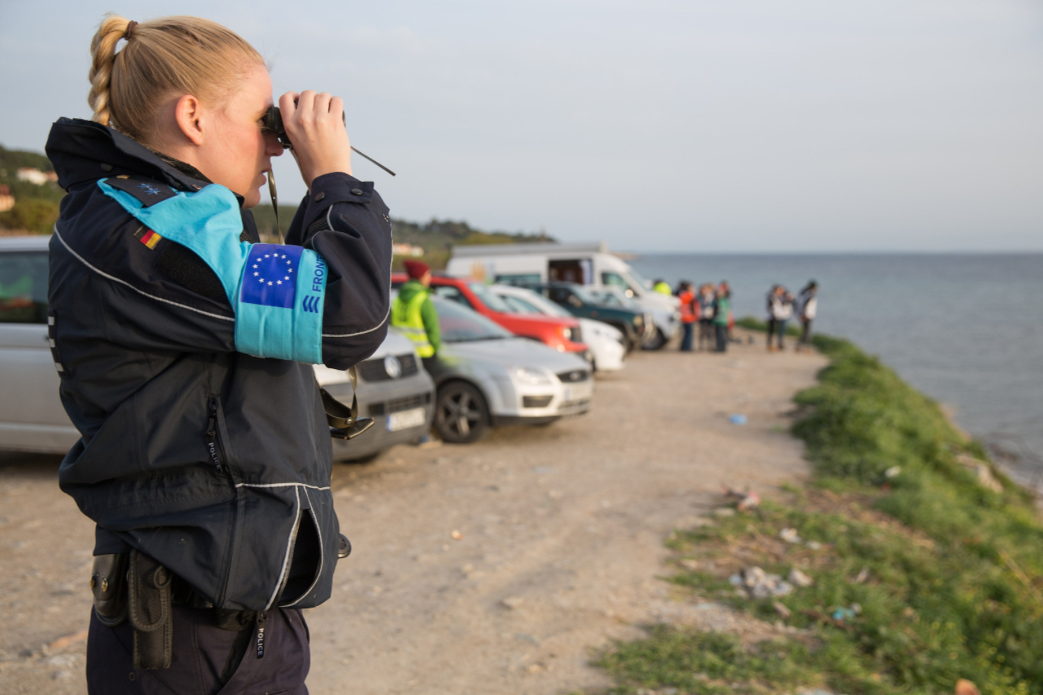 A German federal policewoman wearing a Frontex armband stands on the coast and looks out to sea through her binoculars.