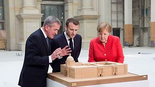 Neil MacGregor, directeur du Forum Humboldt de Berlin, explique à la chancelière fédérale Angela Merkel et au président français Emmanuel Macron le modèle réduit du Forum Humboldt