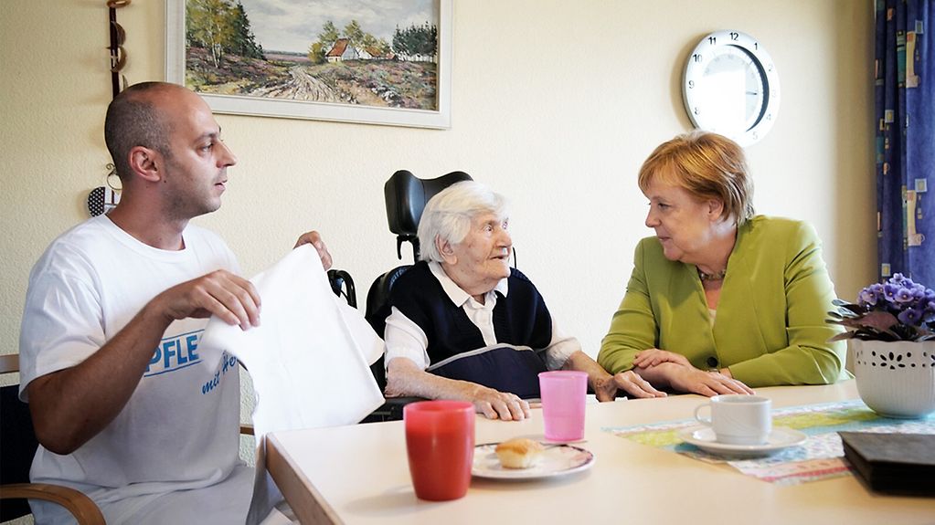 Bundeskanzlerin Angela Merkel mit Pfleger Ferdi Cebi bei einer Bewohnerin des Johannisstift.