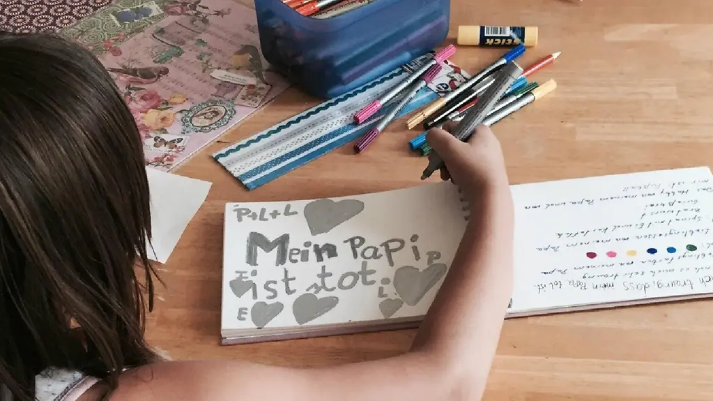 Der Hinterkopf eine dunkelhaarigen Mädchens, das in ein Album malt. Neben vielen grauen Herzchen stehen die Worte "Mein Papi ist tot".
