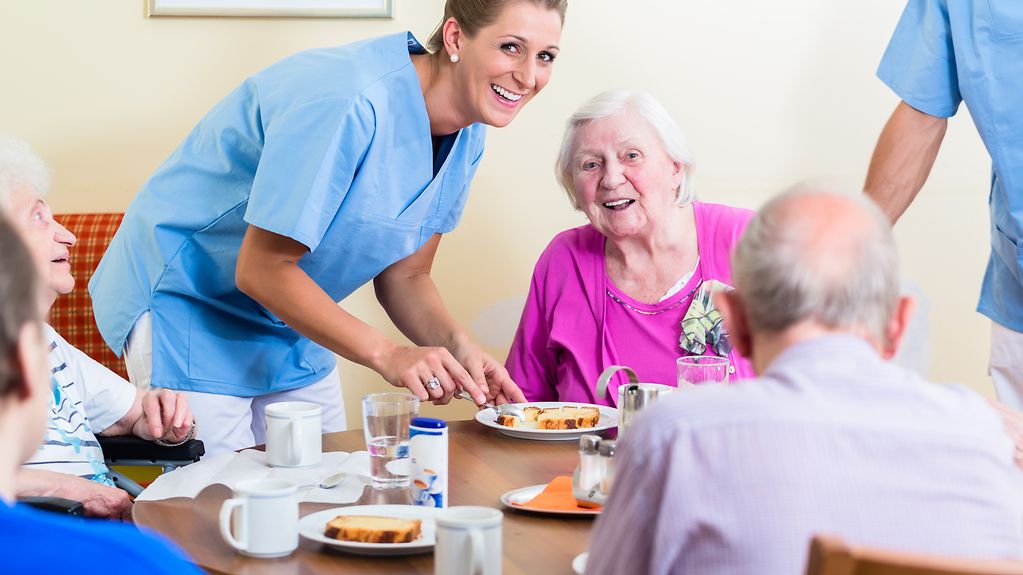 Seniorinnen und Senioren sitzen an einem Tisch beim Essen, eine Pflegerin lächelt freundlich in die Kamera.