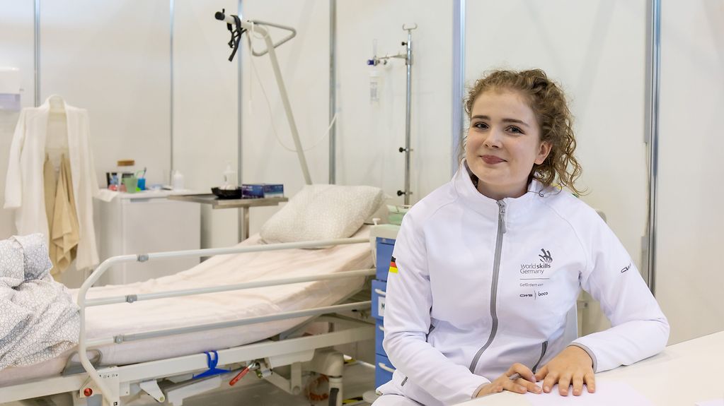 Elisabeth Hölscher sitzt in weißer Kleidung an einem Tisch, im Hintergrund ein Krankenhausbett