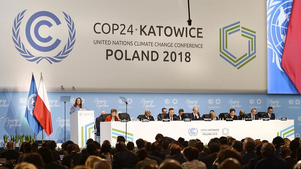 Über die Köpfe einer Menschenmenge hinweg ist ein mit mehreren Personen besetztes Podium zu sehen, darauf die Flaggen der UN und Polens. Auf einem großen Schild dahinter steht "COP24 Katowice - United Nations Climate Change Conference - Poland 2018"