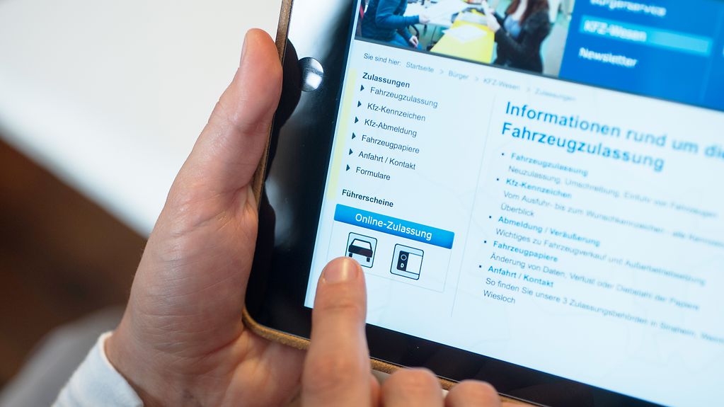 Eine linke Hand hält ein iPad, auf dem die Webseite einer Kfz-Zulassungsstelle geöffnet ist. Die rechte Hand tippt auf eine Stelle auf dem Bildschirm, auf der "Online-Zulassung" zu sehen ist.