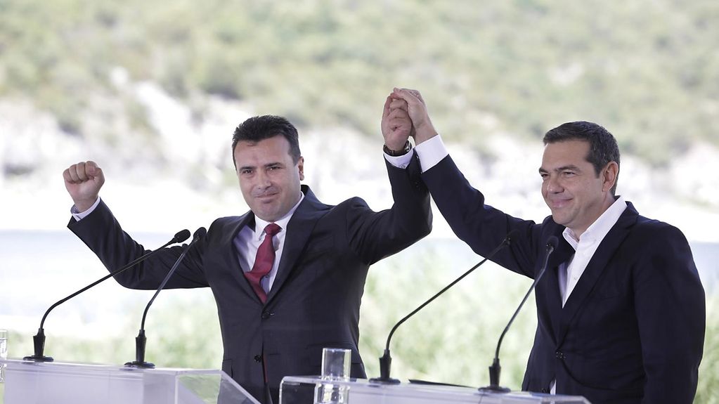 Der griecihische Premierminister Tsipras und der Premierminister der ehemaligen Republik Mazedonien, Zaev, halten sich an den Händen und reißen die Arme in einer Siegergeste hoch. Vor ihnen Stehpulte. 