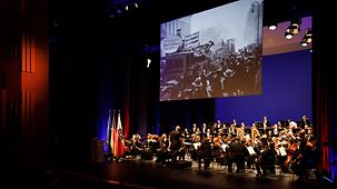 Blick auf die Bühne beim Festakt mit Orchester und historischem Film auf einer Leinwand.