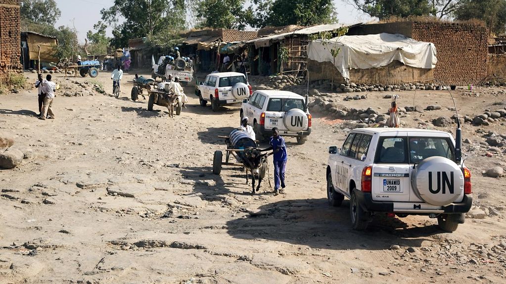 Jeeps mit UN-Zeichen fahren in einer Kolonne eine staubige, unebene Straße entlang. Daneben laufen vereinzelt Sudaneser mit Karren.