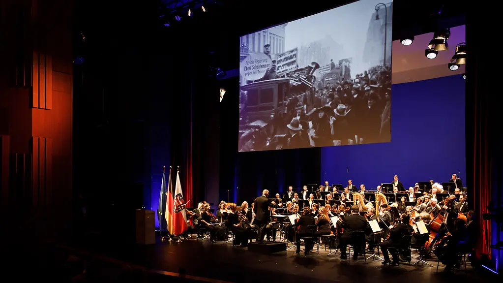 A view of the stage; an orchestra plays while a film from the period is shown on a screen.