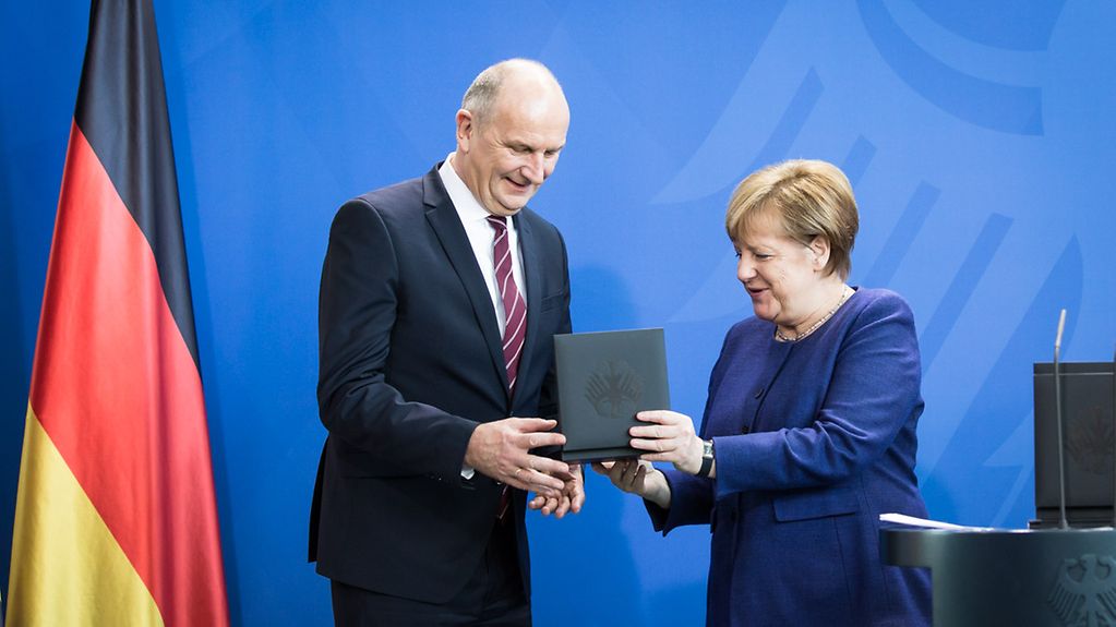 Die Kanzlerin übergibt dem brandenburgischen Ministerpräsidenten Dietmar Woidke die Gedenkmünze in einer dunkelfarbigen Box mit dem Bundesadler, dem deutschen Wappen, auf dem Deckel. Der Deckel ist geöffnet, damit Woidke die Münze inspizieren kann.