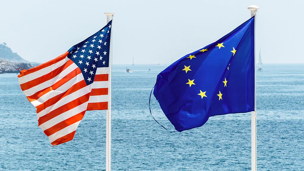 Die Flaggen der Europäischen Union und der USA wehen im Vordergrund, im Hintergrund der Ozean.