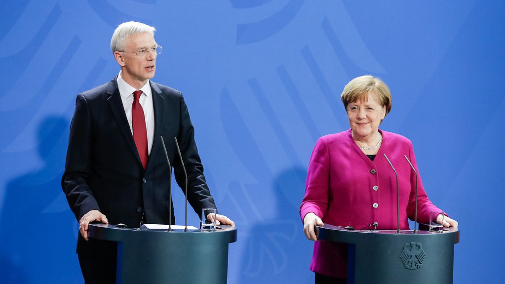 Against a blue backdrop, Chancellor Angela Merkel and Latvian Prime Minister Krišjānis Kariņš each stand at a lectern during the press conference.