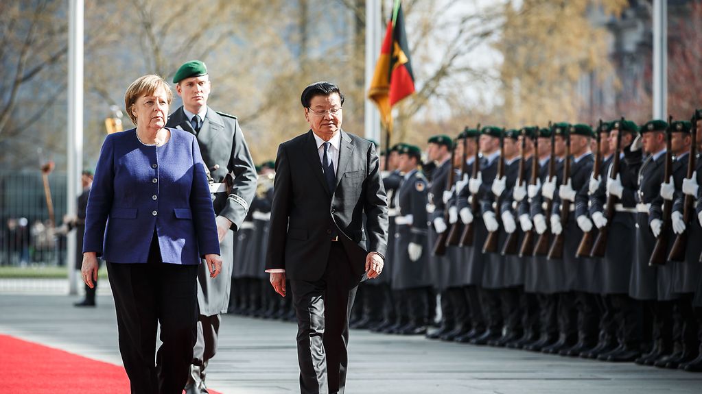 Auf einem roten Teppich laufen Kanzlerin Merkel und der laotische Premierminister an einem Spalier aus Bundeswehrsoldatinnen und Soldaten in Uniform entlang.