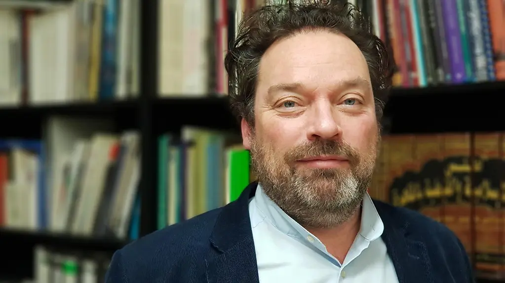 Professor Stefan Weber trägt einen dunkelblaune Sakko und ein weißes Hemd. Er steht vor einer Bücherwand.