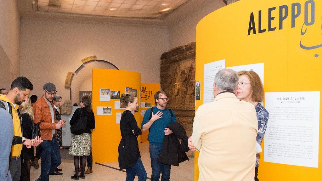 Auf einem gelben Aufsteller steht Aleppo, zu sehen sind menschen bei der Ausstellungseröffnung. Im Hintergrund ein großes archäologisches Fundstück.