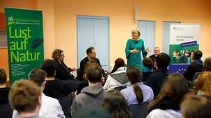The Federal Agriculture Minister Julia Klöckner stands at the front of a classroom beside a large green poster bearing the message "Lust auf Natur" (enjoy nature) and talks to students.