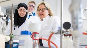 Federal Environment Minister Svenja Schulze performs an experiment in a laboratory of the Hans-Böckler-Berufskolleg with a student wearing a headscarf and a blond student.