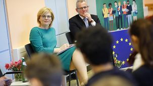 Julia Klöckner and a grey-haired gentleman sit in a classroom next to an EU flag during a discussion with students.