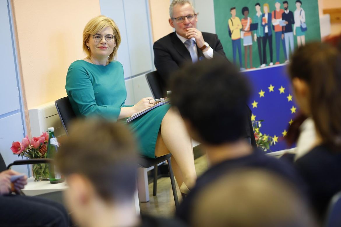 Julia Klöckner and a grey-haired gentleman sit in a classroom next to an EU flag during a discussion with students.