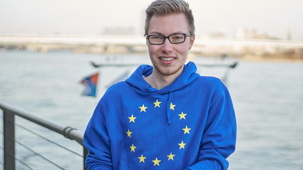 Hannich, ein junger Mann mit blonden Haaren, schwarz-umrandeter Brille und blauem Kapuzenpullover mit gelben Europa-Sternen drauf, lehnt lässig an einem Metallgeländer am Hafen.