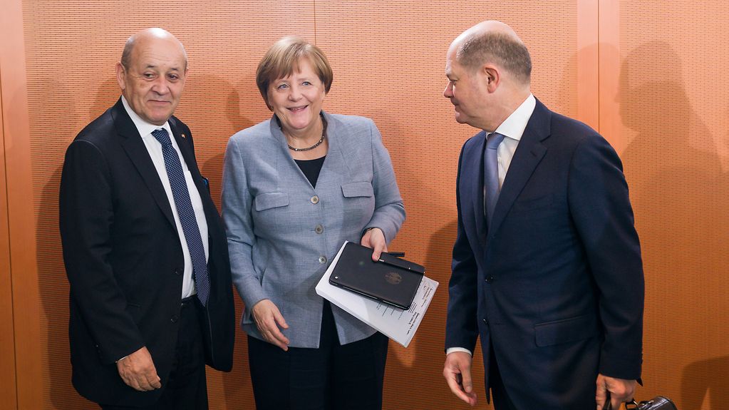 Frankreichs Außenminister Le Drian, Kanzlerin Merkel und Vizekanzler Scholz stehen vor einer holzvertäfelten Wand. 