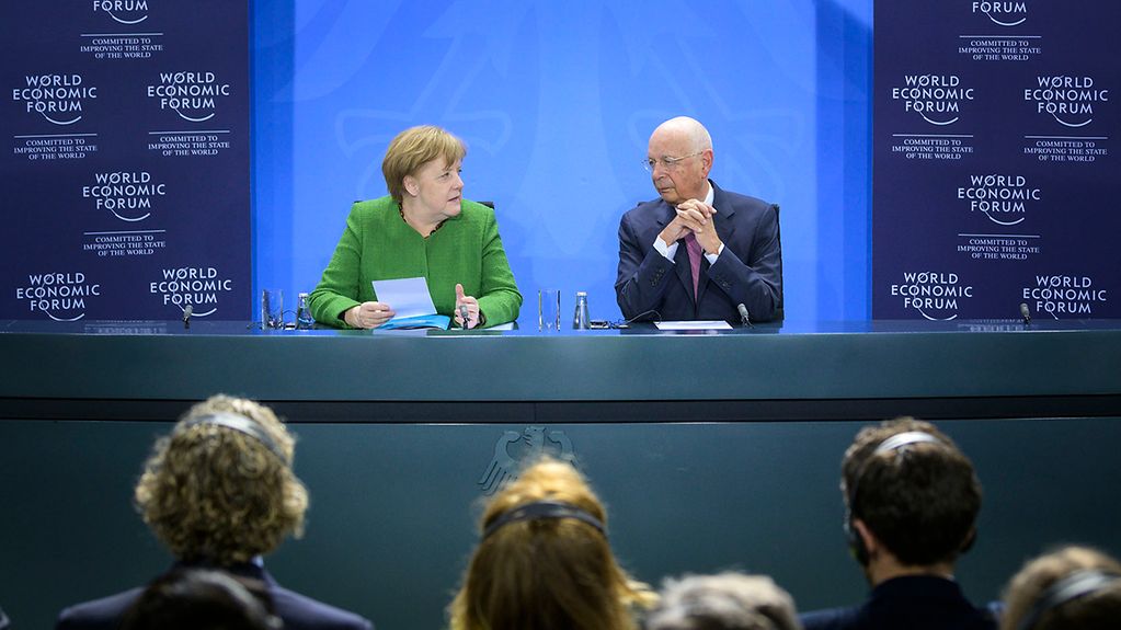 Kanzlerin Merkel und WEF-Vorsitzender Schwab sitzen an einem Rednertisch vor einer blauen Wand. Im Vordergrund sind unscharf die Köpfe der Zuhörer zu erkennen.
