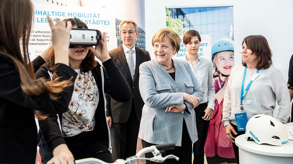 Roberta sitzt auf einem Fahrrad im Kanzleramt. Sie trägt eine VR-Brille, die ihre Freundin ihr gerade aufsitzt. Merkel und andere schauen interessiert zu.