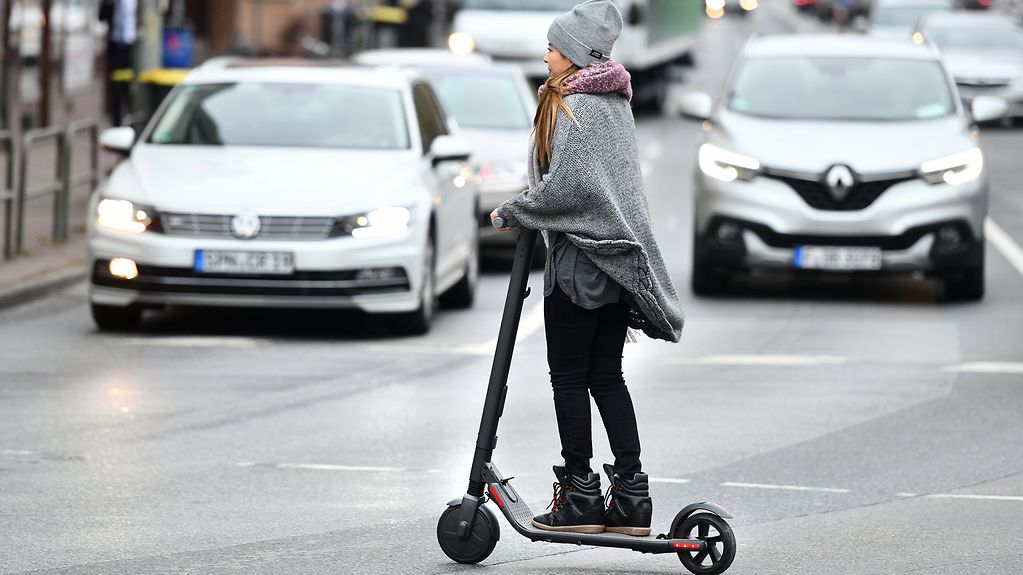 Eine junge Frau mit grauer stylischer Wollmütze fährt mit ihrem elektrischen Tretroller in der Stadt und überquert auf dem Roller die Straße.