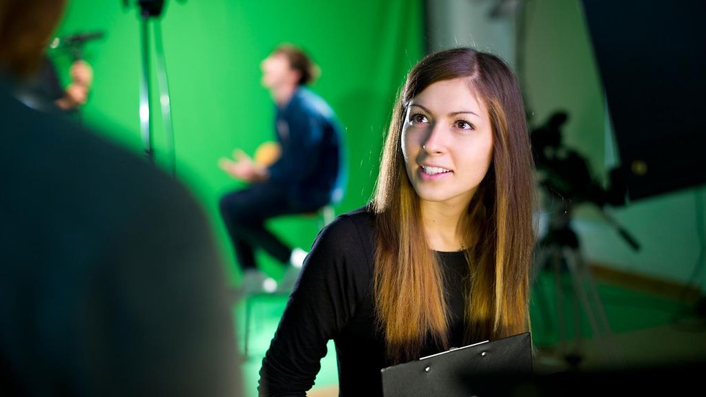 Auszubildende in einem Fernsehstudio. Im Hintergrund unscharf zu erkennen ist ein Mann im Scheinwerferlicht, der in eine Kamera spricht.