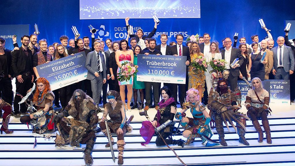Auf einer Bühne vor einer blauen Wand mit dem Schriftzug "Deutscher Computerspielpreis 2019" posieren die Sieger des Deutschen Computerspielpreises, die Minister Bär und Scheuer sowie als Spielfiguren verkleidete Leute und jubeln.