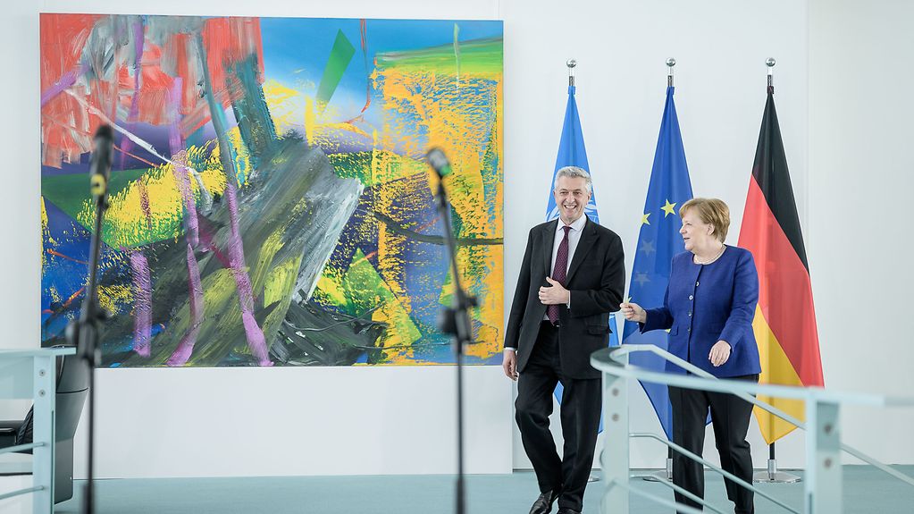 Kanzlerin Merkel und UN-Flüchtlinskommissar Grandi gehen den Gang im Kanzleramt entlang. Hinter ihnen zu sehen sind die Flaggen Deutschlands, der EU und der UNO, dazu ein buntes Wandgemälde.