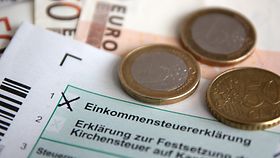 Antrag zur Einkommenssteuererklärung im Ausschnitt. Auf dem Antrag liegen einige Euro-Münzen. Im Hintergrund finden sich drei 50-Euro-Scheine als Fächer gelegt. 
