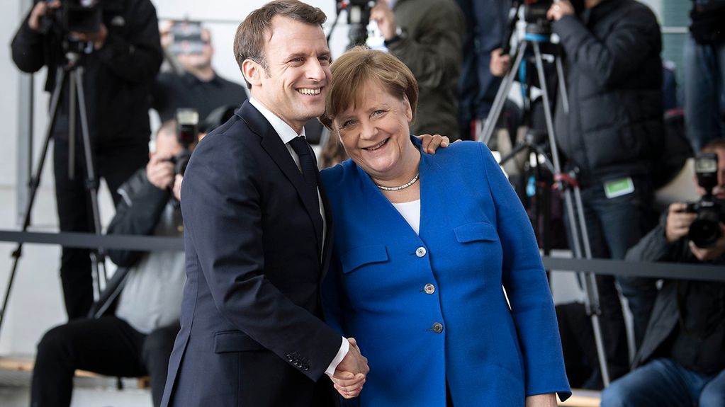 Umgeben von Kameras und Stativen schütteln sich Kanzlerin Merkel und der französische Präsident Macron die Hand und lächeln dabei fröhlich.