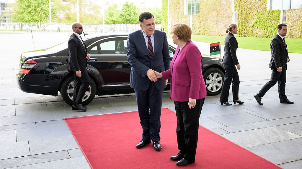 Debout sur le tapis rouge, Mme Merkel et Fayez al-Sarraj se serrent la main. La chancelière est placée de profil et tourne la tête à la caméra. En arrière-plan, une limousine