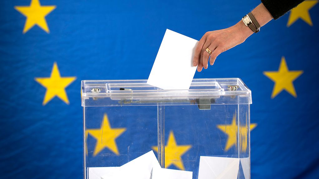 Vor einer großen Europaflagge auf blauem Grund mit gelben Sternen steht eine durchsichtige Plastikbox, in der sich bereits Umschläge befinden und eine Hand einen weiteren Umschlag hineinwirft.