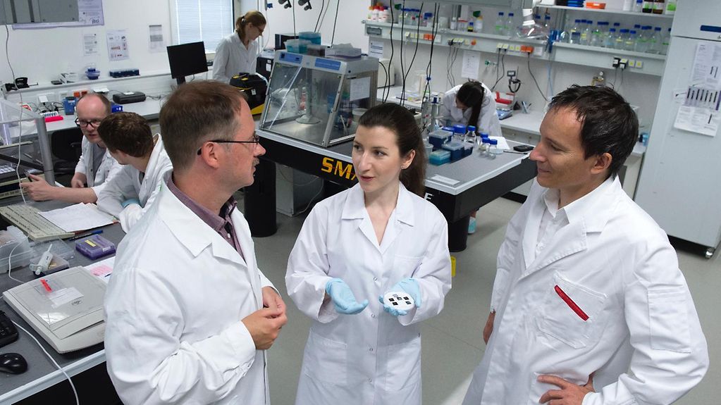 Drei Forscher stehen in weißen Kitteln in einem Labor und unterhalten sich. Im Hintergrund sind weitere Forscher bei der Arbeit zu sehen.