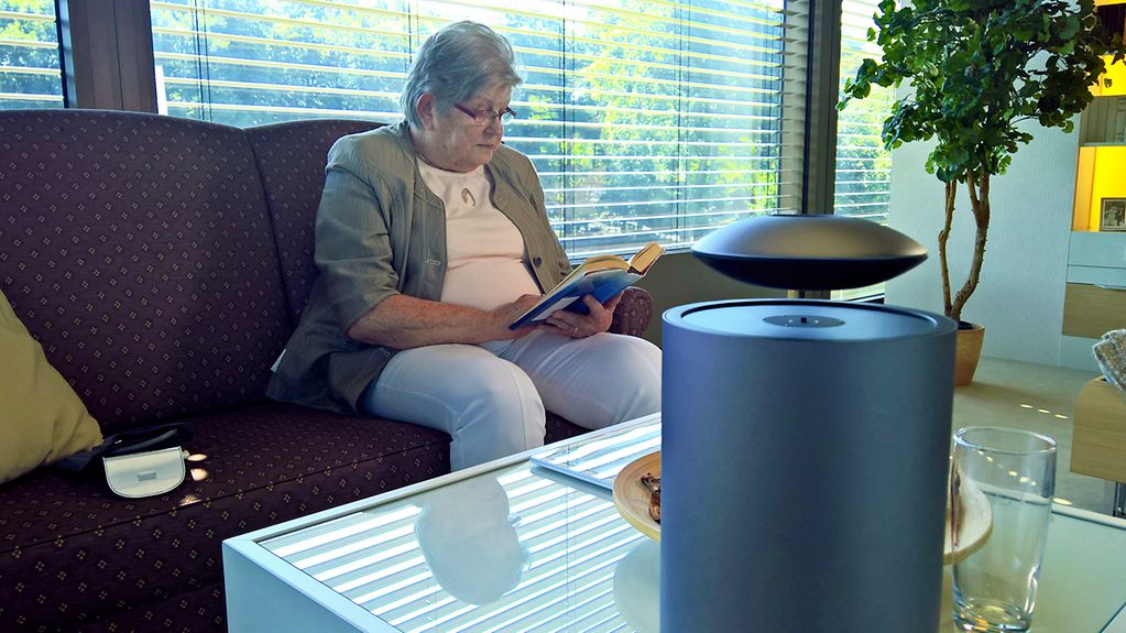 Eine ältere Dame sitzt auf einem Sofa und liest ein Buch. Im Vordergrund steht ein intelligenter Sprachassistent in Form einer Lautsprecherbox auf dem Couchtisch.