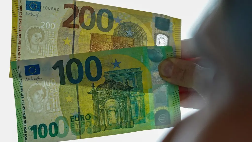 Eine nur unscharf zu erkennene Person hält einen 100- und einen 200-Euro-Schein gegen das Licht. Die Scheine sind zu einem Fächer gelegt.