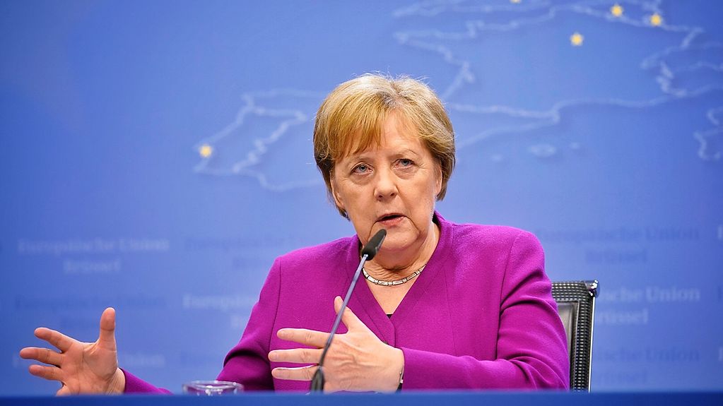 Chancellor Merkel gesticulates as she speaks into a microphone on the table in front of her. She wears a pink blazer and sits in front of a blue background.