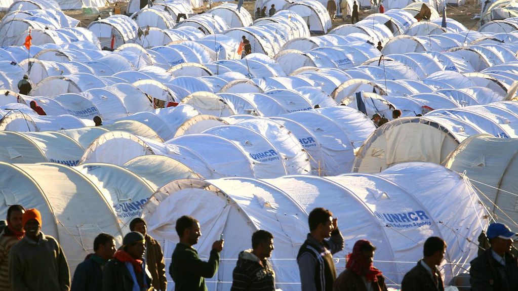 Flüchtlinge stehen in einer Schlange, im Hiintergrund ein aus weißen Zelten bestehendes Flüchtlingscamp.