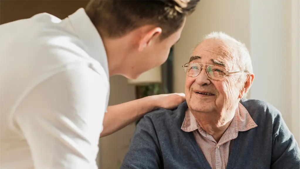 An elderly man sits and looks up at a young nurse in a white coat, who is leaning down to him and touching his shoulder.