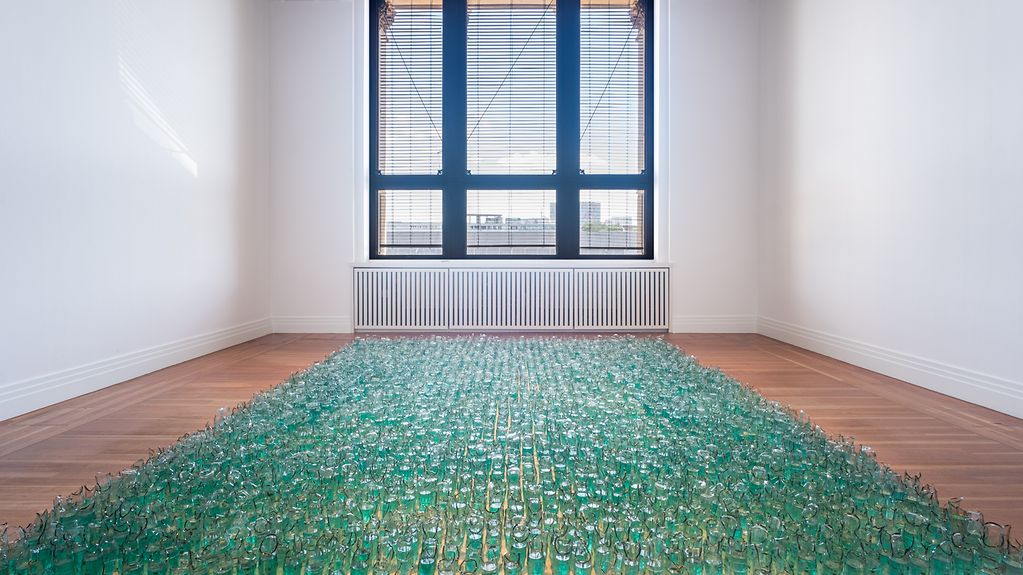 Mit grüner Flüssigkeit befüllte Flaschen stehen als Rasenfläche angeordnet im Raum.