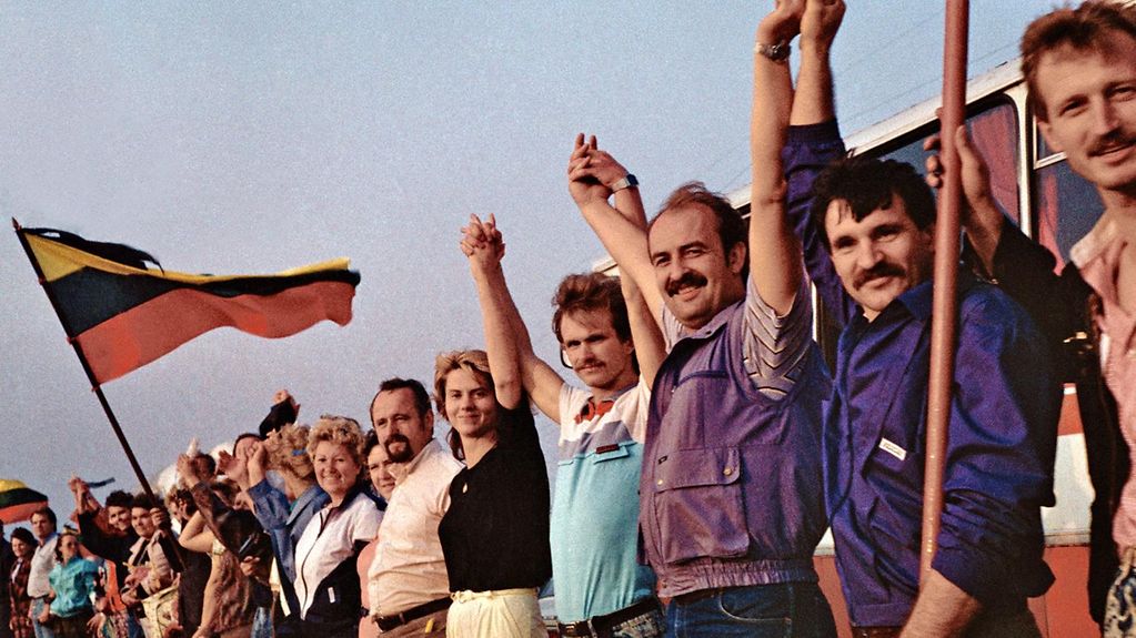 Männer und Frauen halten sich an den Händen und bilden eine Menschenkette. Im Hintergrund weht die litautische Nationalflagge. 