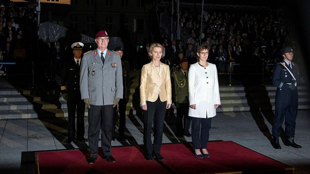 Ein Soldat in Uniform, Ministerin a.D. von der Leyen und Verteidigungsministerin Kramp-Karrenbauer auf einem roten Teppich bei Nacht. Im Hintergrund Soldaten.